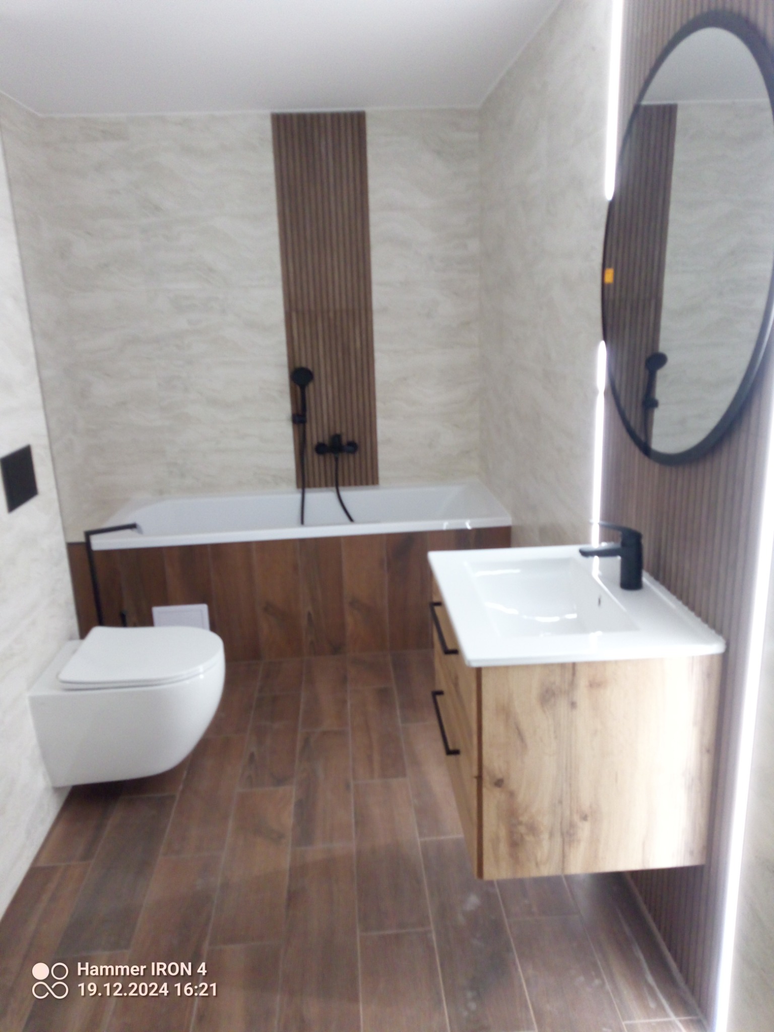 Nowoczesna łazienka z drewnianymi akcentami: wisząca toaleta, umywalka z szafką i okrągłe lustro. Beżowe płytki imitujące marmur na ścianach i brązowe płytki na podłodze.