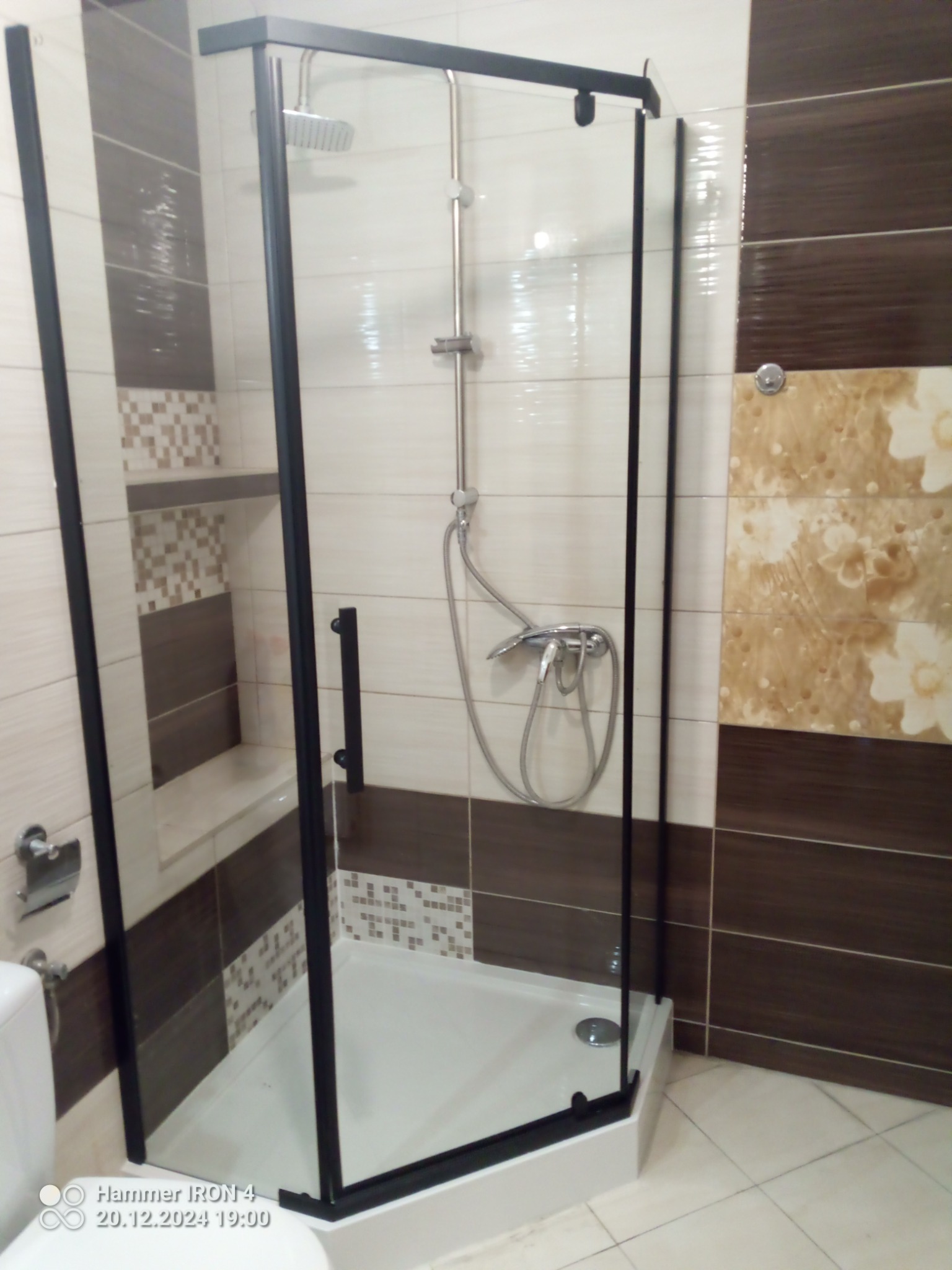 Nowoczesna kabina prysznicowa z czarnymi profilami, jasne płytki z dekorami w beżach i brązach. Widoczny zestaw prysznicowy i fragment toalety.