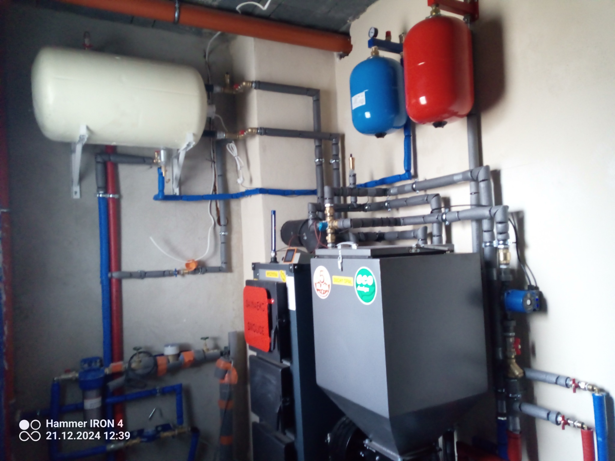 Złożona instalacja grzewcza z piecem na paliwo stałe, zbiornikami wyrównawczymi (czerwony i niebieski) oraz naczyniem przeponowym (biały). Widoczne rury i zawory.