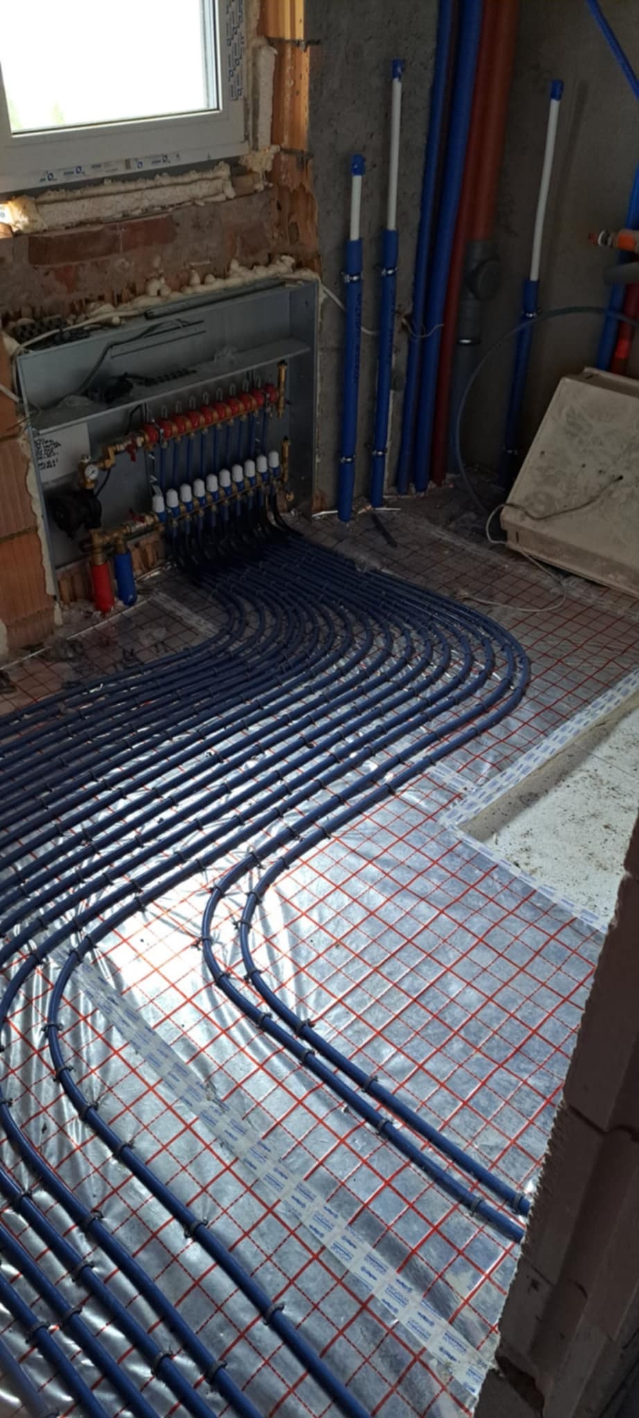 Niebieskie rury ogrzewania podłogowego ułożone na folii z czerwoną siatką, widoczne elementy rozdzielacza i piony instalacyjne przy ścianie w surowym stanie.
