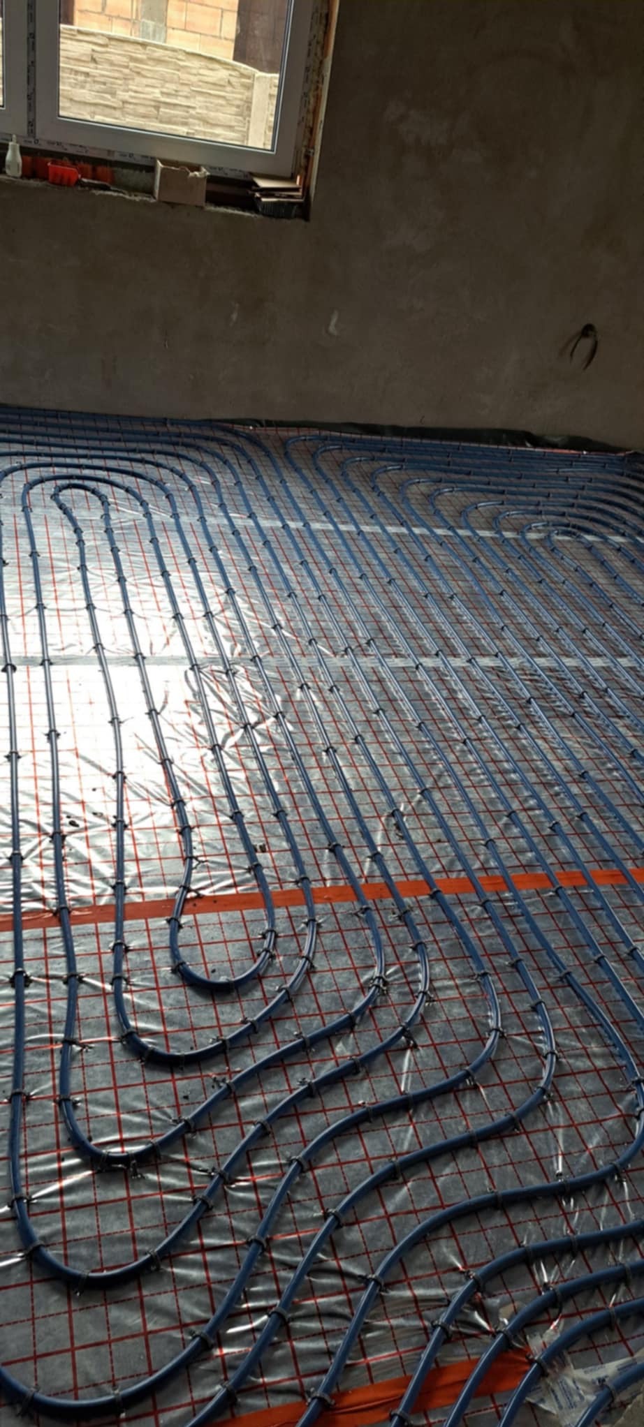 Ułożona instalacja ogrzewania podłogowego z niebieskich rurek na folii z czerwoną siatką, widok z góry, surowy stan wykończenia wnętrza.