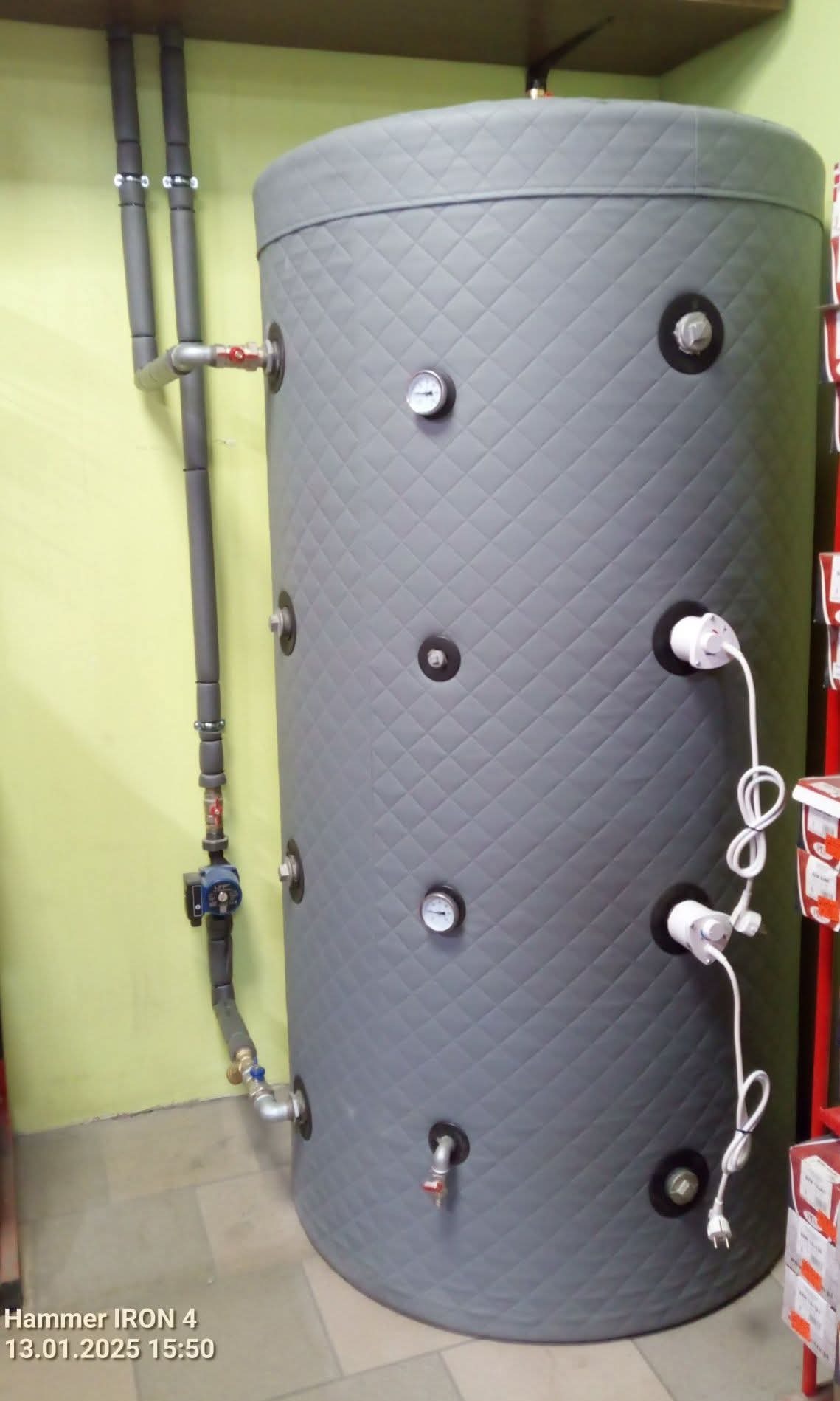 Szary, ocieplony zasobnik ciepłej wody użytkowej z podłączonymi rurami i elementami instalacji, widoczne wtyczki elektryczne.