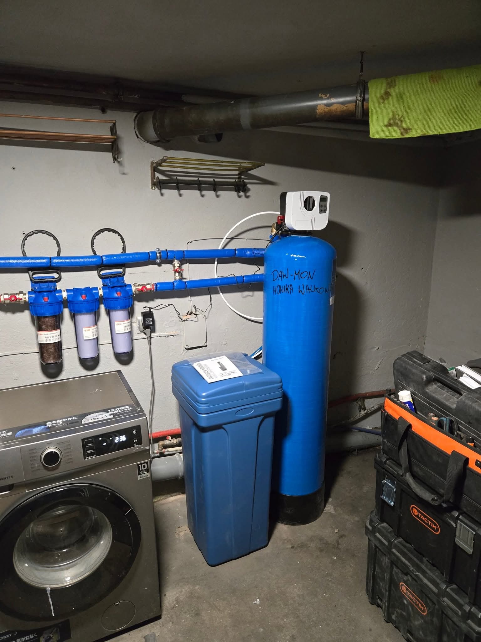 Instalacja uzdatniania wody w piwnicy obok pralki, z niebieskimi filtrami i zbiornikiem. Widoczne rury, skrzynki narzędziowe i fragment suszarki ściennej.