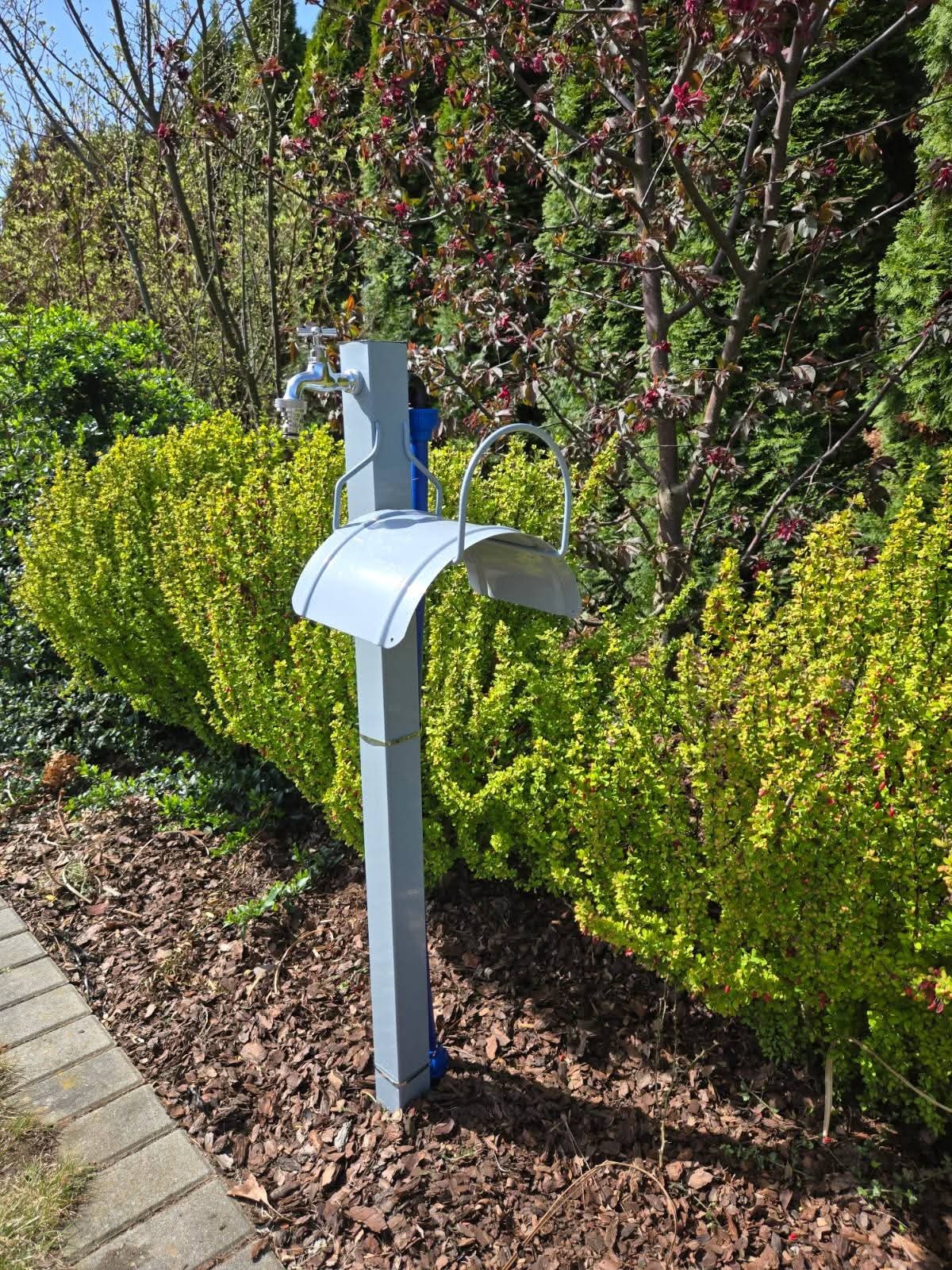 Zewnętrzny, szary kran ogrodowy z osłoną, umieszczony na trawniku z krzewami i drzewami. Kran zamontowany na metalowym słupku. Widok na ogród.