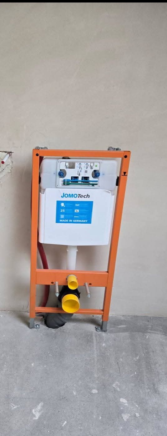 Pomarańczowa rama podtynkowa JomoTech z podłączonymi rurami kanalizacyjnymi, zamontowana przy surowej ścianie w trakcie prac hydraulicznych.