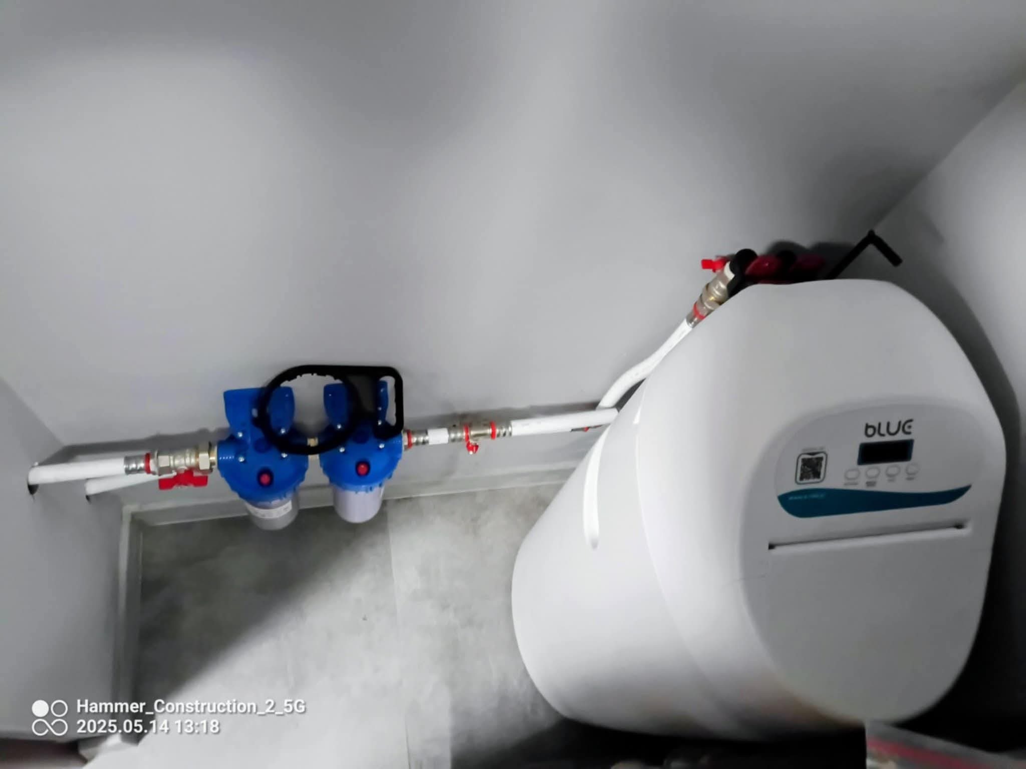 Biały bojler elektryczny marki Blue, zamontowany na ścianie z filtrem wody, połączony rurami. Widok z dołu, oświetlenie sufitowe.