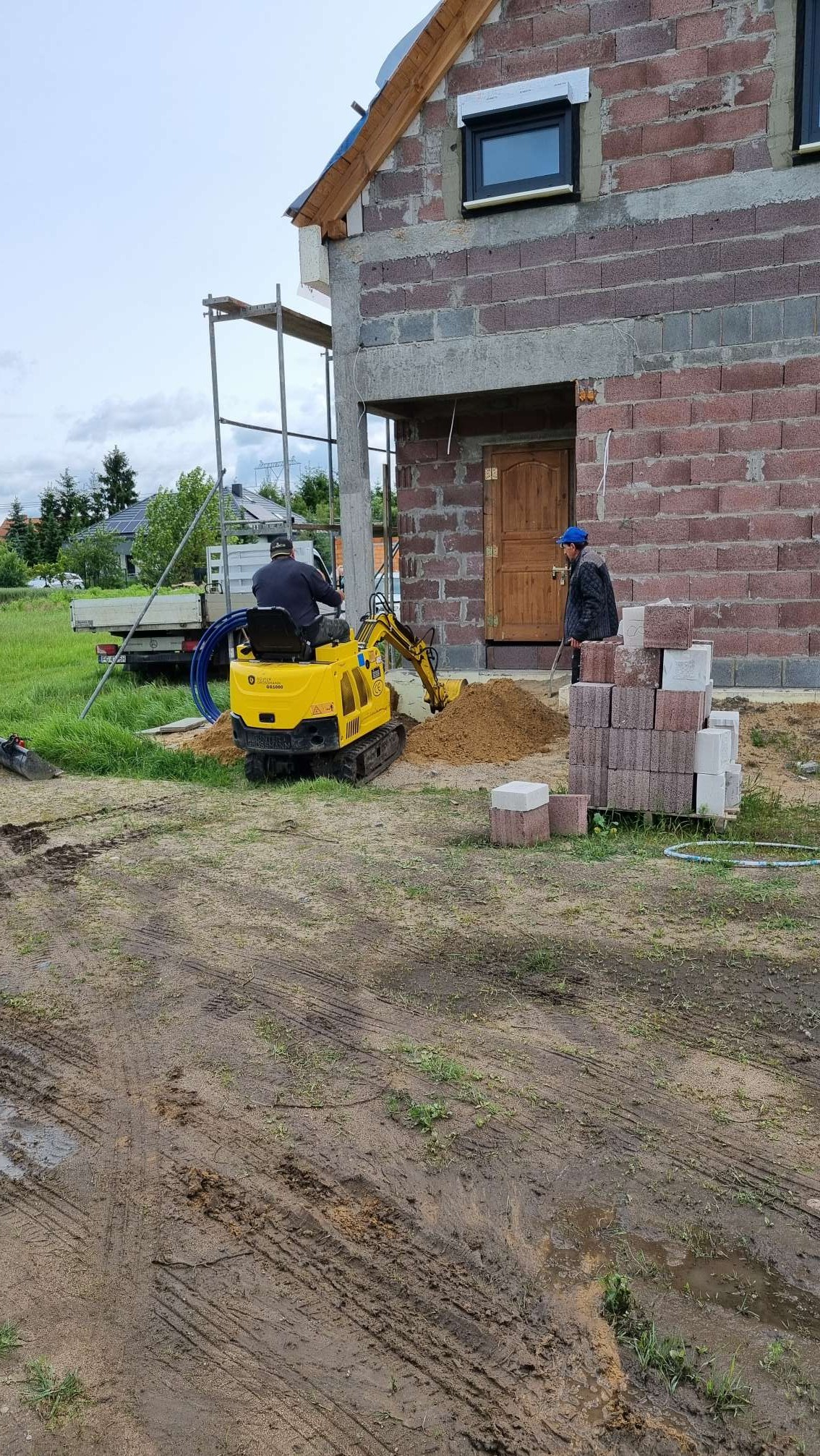 Żółta minikoparka przy budynku w trakcie budowy, wykopuje ziemię. Obok sterta bloczków betonowych i pracownik w niebieskiej czapce.