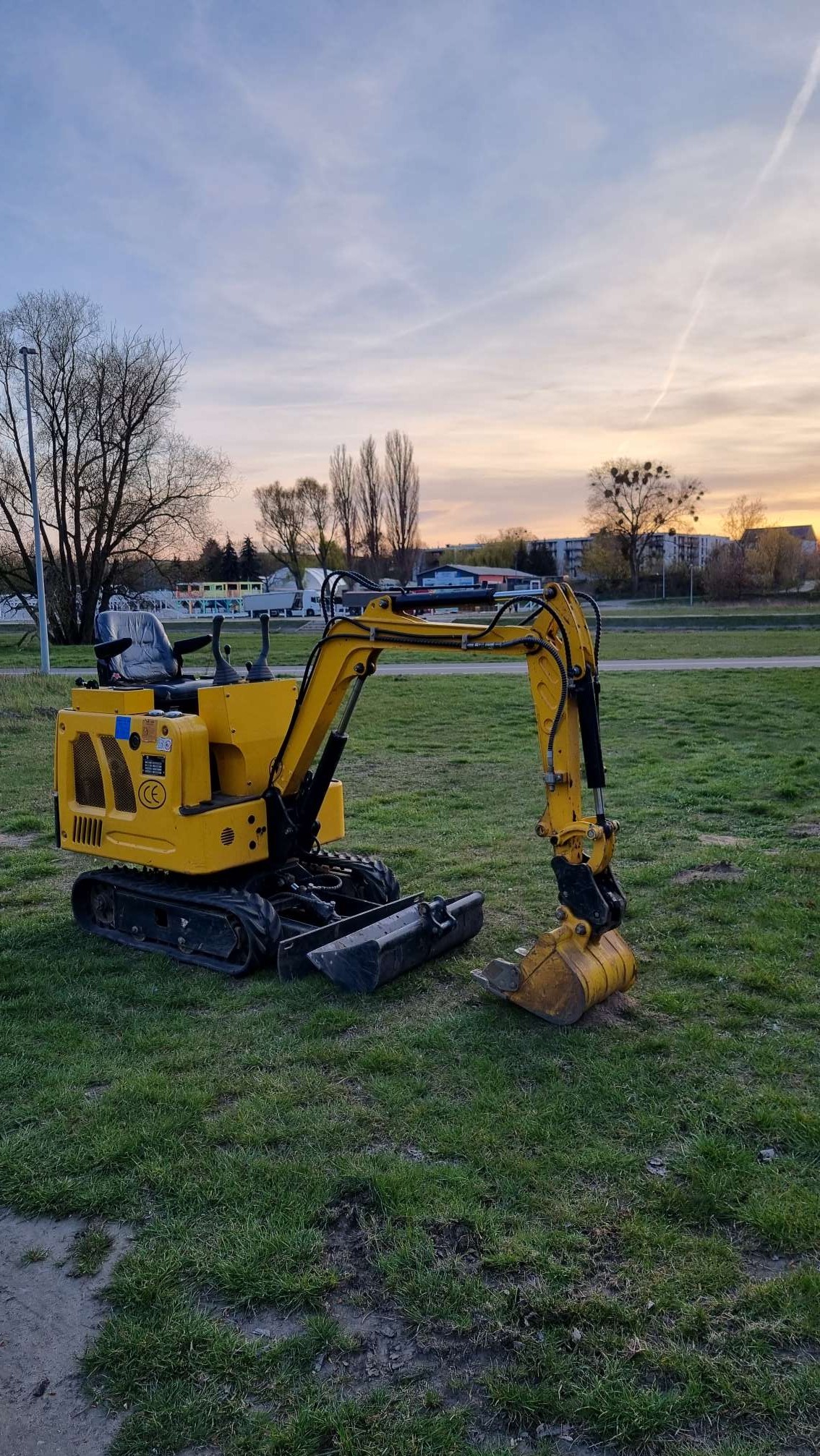 Żółta minikoparka na trawie, widok z boku. Na drugim planie drzewa i budynki pod wieczornym niebem. Sprzęt budowlany gotowy do pracy.