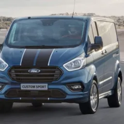 Niebieski Ford Transit Custom Sport z czarnymi pasami na masce, dynamicznie prezentowany na torze wyścigowym. Widoczne detale: logo Forda i oznaczenie modelu.