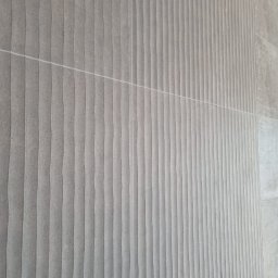 KJP Wykończenia wnętrz Tomasz Pietras - Ściana wyłożona szarymi, strukturalnymi płytkami z delikatnym wzorem fal, ułożonymi w pionowe pasy, z widocznymi fugami. Minimalistyczny design.