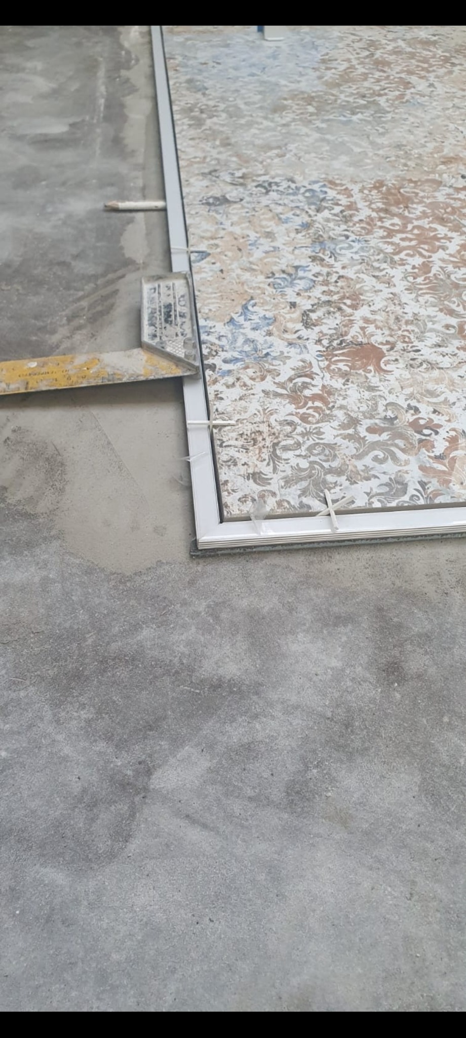 Układanie dekoracyjnej płytki podłogowej z użyciem listew i poziomicy, widoczny fragment surowego podłoża, etap prac glazurniczych.