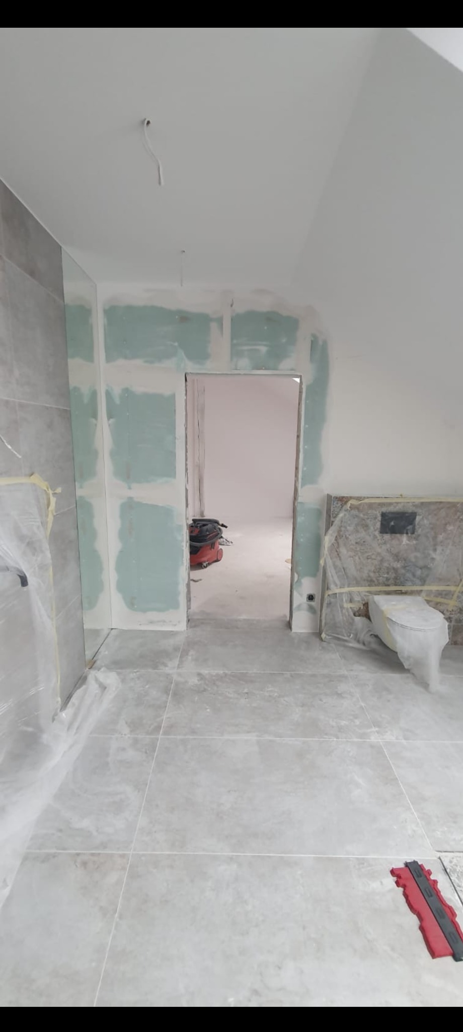 Łazienka w trakcie prac: szare płytki podłogowe, częściowo położone płytki ścienne, płyty karton-gips na ścianach, zabezpieczona toaleta folią. Widoczny sprzęt budowlany.
