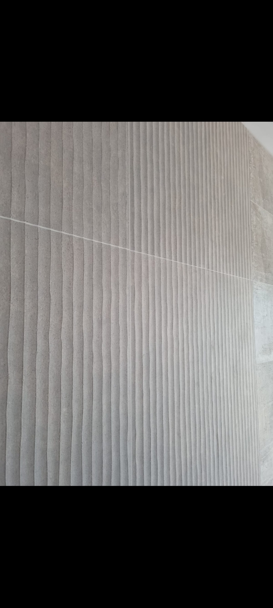 Ściana wyłożona szarymi, strukturalnymi płytkami z delikatnym wzorem fal, ułożonymi w pionowe pasy, z widocznymi fugami. Minimalistyczny design.