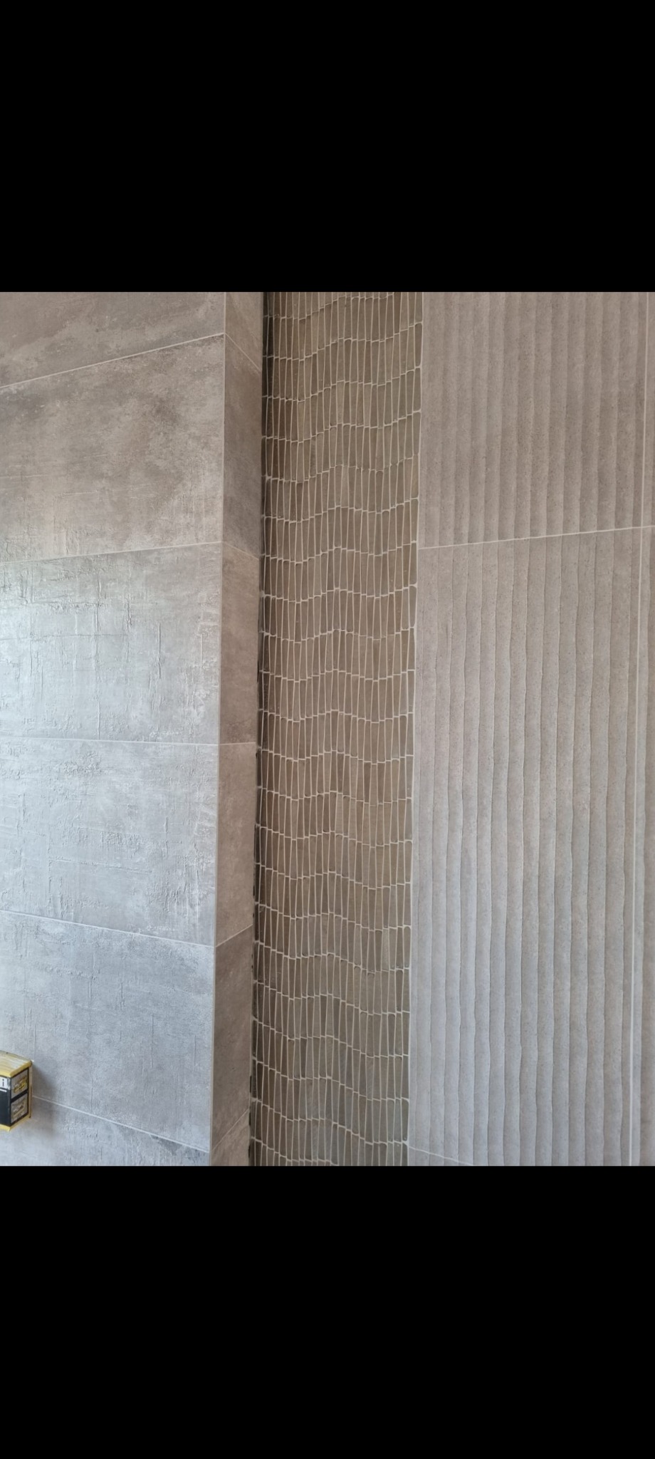 Ściana wykończona szarą, strukturalną glazurą z dekoracyjnym pasem mozaiki. Widoczny fragment narożnika i poziomych fug. Realizacja w łazience.