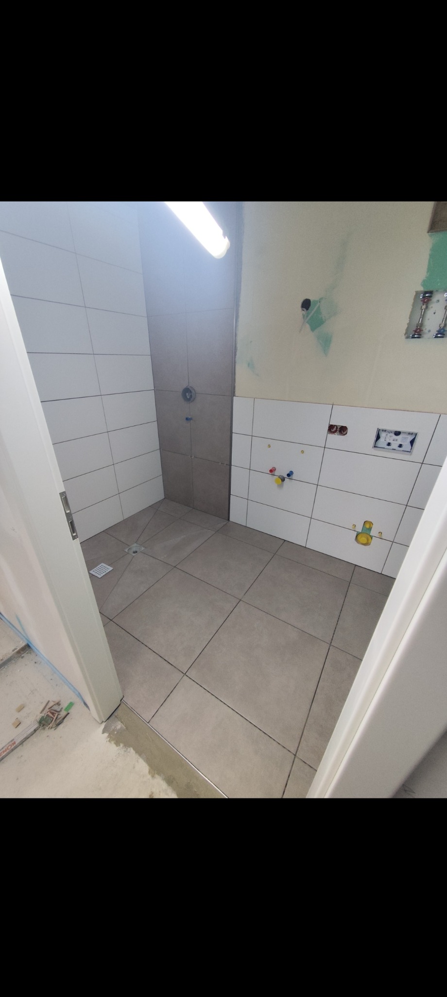 Łazienka w trakcie remontu: ułożone płytki podłogowe i ścienne, widoczne instalacje, białe drzwi, szare płytki w strefie prysznica. Częściowo wykończone ściany.