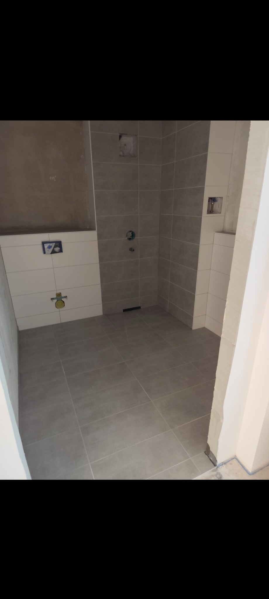 Wykończona łazienka w szarościach z częściowo położonymi białymi płytkami. Widoczne instalacje pod umywalkę i baterię. Minimalistyczny design.