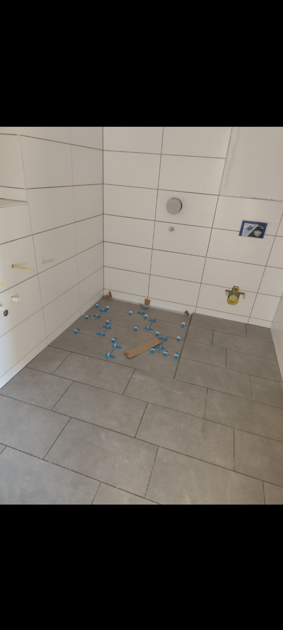 Szare płytki podłogowe w trakcie układania w łazience. Widoczne krzyżyki dystansowe i fragment niezakończonej powierzchni. Białe płytki ścienne w tle.