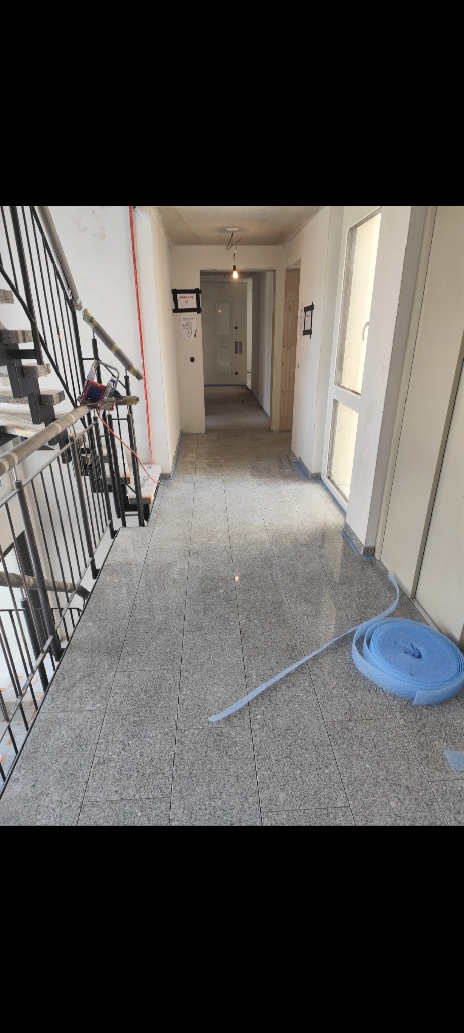 Szare płytki podłogowe w korytarzu budynku, zabezpieczone niebieską folią ochronną przy ścianach i drzwiach. Widoczne schody i narzędzia glazurnicze.