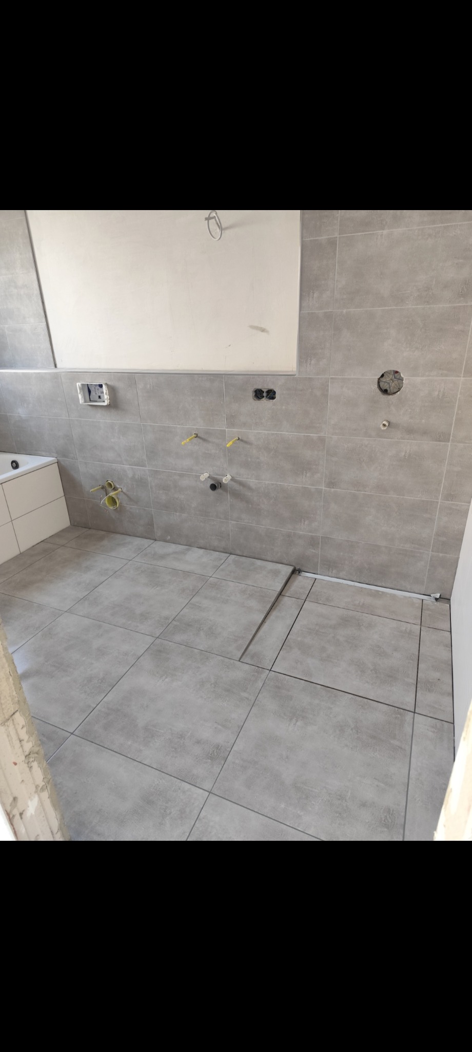 Łazienka w trakcie remontu: szare płytki na ścianach i podłodze, widoczne instalacje, fragment wanny. Ujęcie z niskiego kąta.