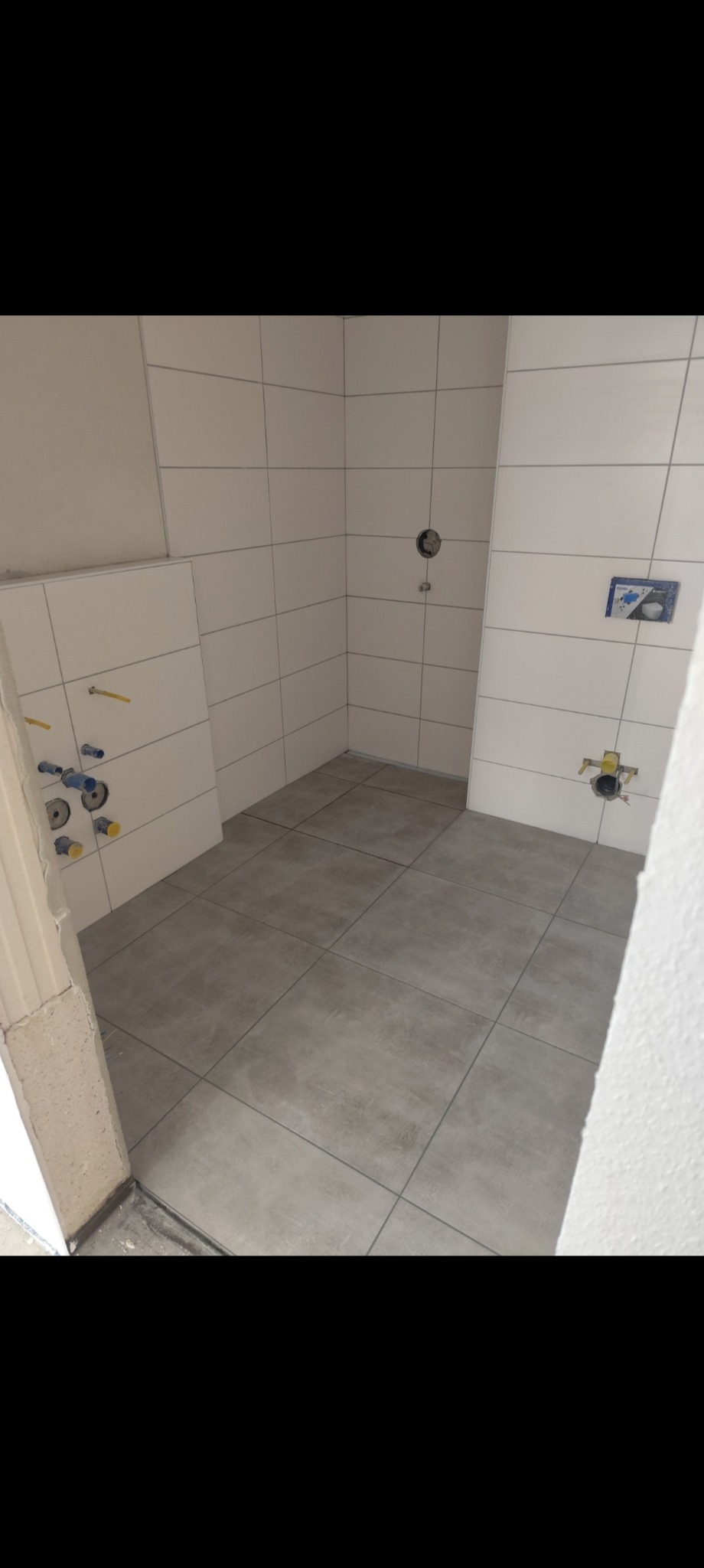 Wykończona łazienka w jasnych płytkach z szarą podłogą. Widoczne przyłącza wodne i kanalizacyjne na ścianach, przygotowane do montażu armatury. Minimalistyczny styl.