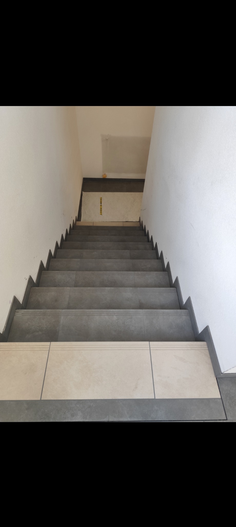 Wykończone schody z szarymi i beżowymi płytkami, widok z góry. Szare płytki na stopniach, beżowe na podstopnicach, szara listwa wykończeniowa. Białe ściany.