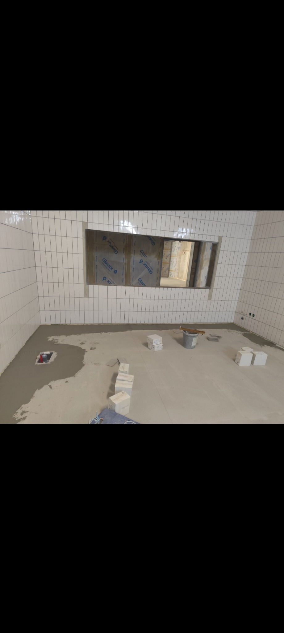 Białe, prostokątne płytki na ścianie łazienki z wnęką. Na podłodze wylewka samopoziomująca, przygotowana do położenia płytek. Widoczne narzędzia i materiały.