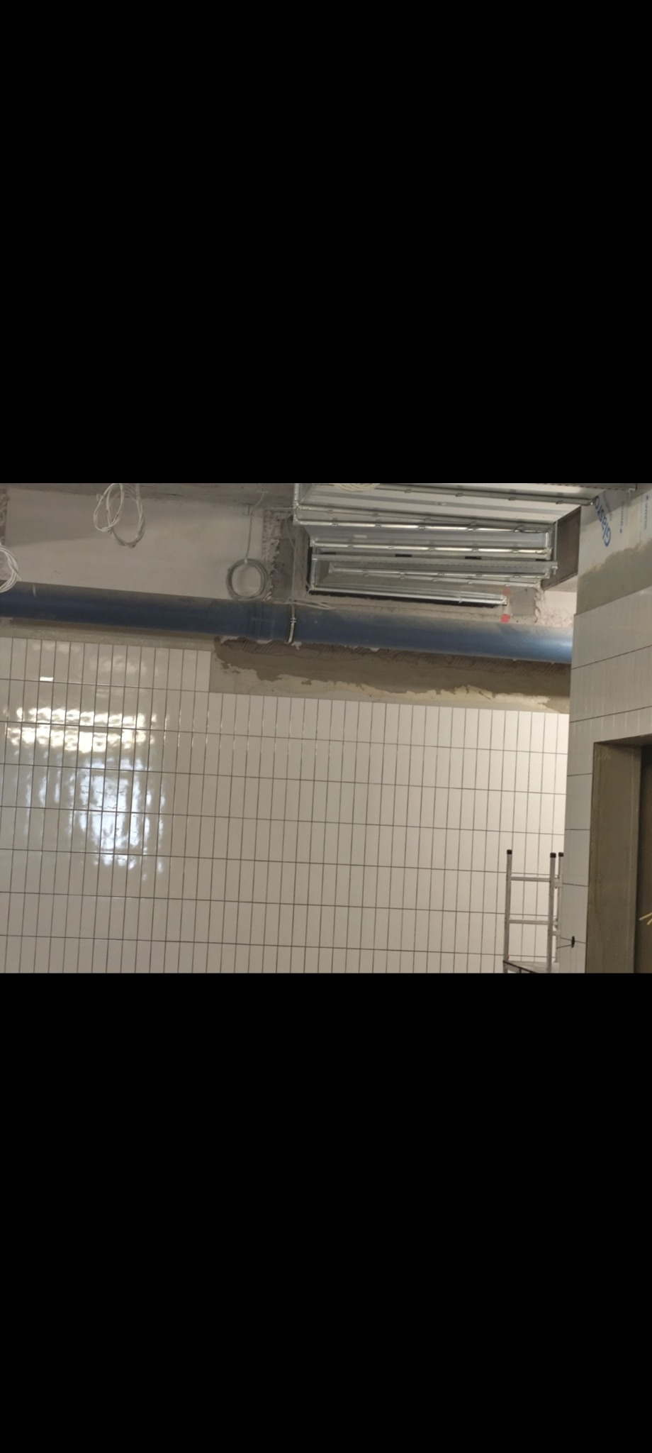 Białe, wąskie płytki ułożone na ścianie, widoczne rury i instalacje pod sufitem, drabina w tle. Prace wykończeniowe w trakcie realizacji.