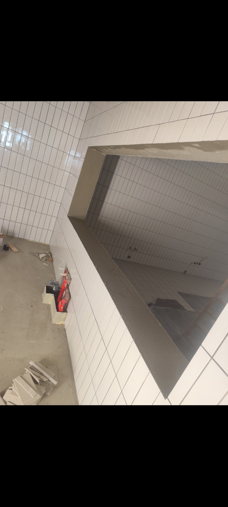 Wnętrze w trakcie prac glazurniczych; białe, prostokątne płytki ułożone na ścianach i podłodze, widoczne materiały budowlane i fragment instalacji.