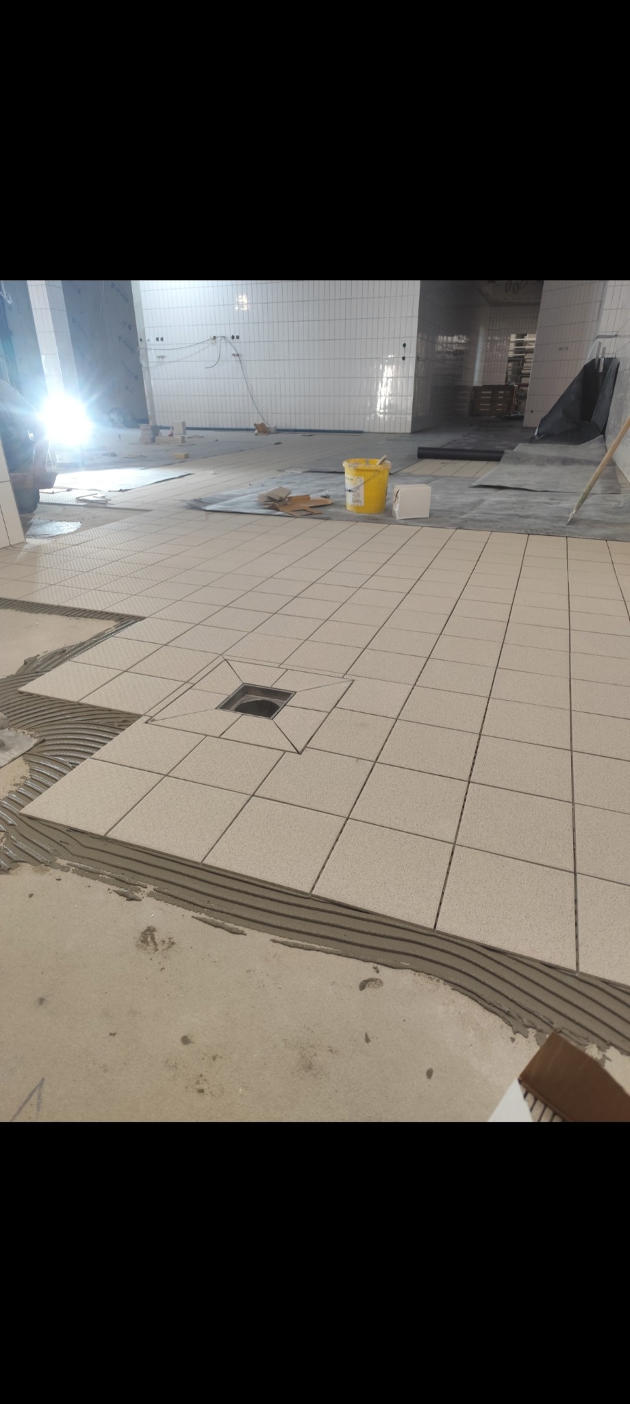 Układanie jasnych płytek podłogowych w dużej przestrzeni, widoczny odpływ liniowy i wiaderko z klejem. Prace glazurnicze w trakcie realizacji.