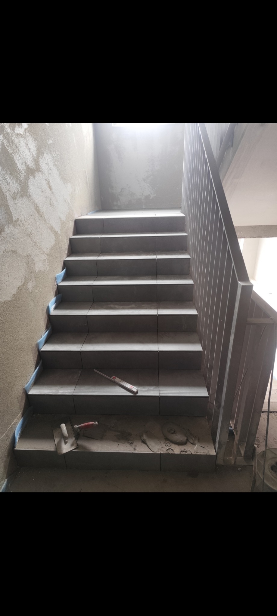 Schody w trakcie układania płytek, widoczne narzędzia glazurnicze i surowe ściany. Ujęcie z dołu schodów, podkreślające etap prac wykończeniowych.