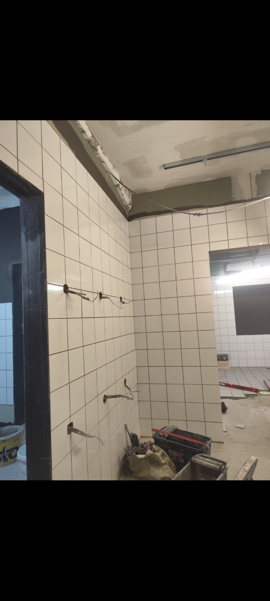 Białe płytki na ścianie w trakcie układania, widoczne przewody elektryczne i narzędzia glazurnicze. Prace w Sycowie.