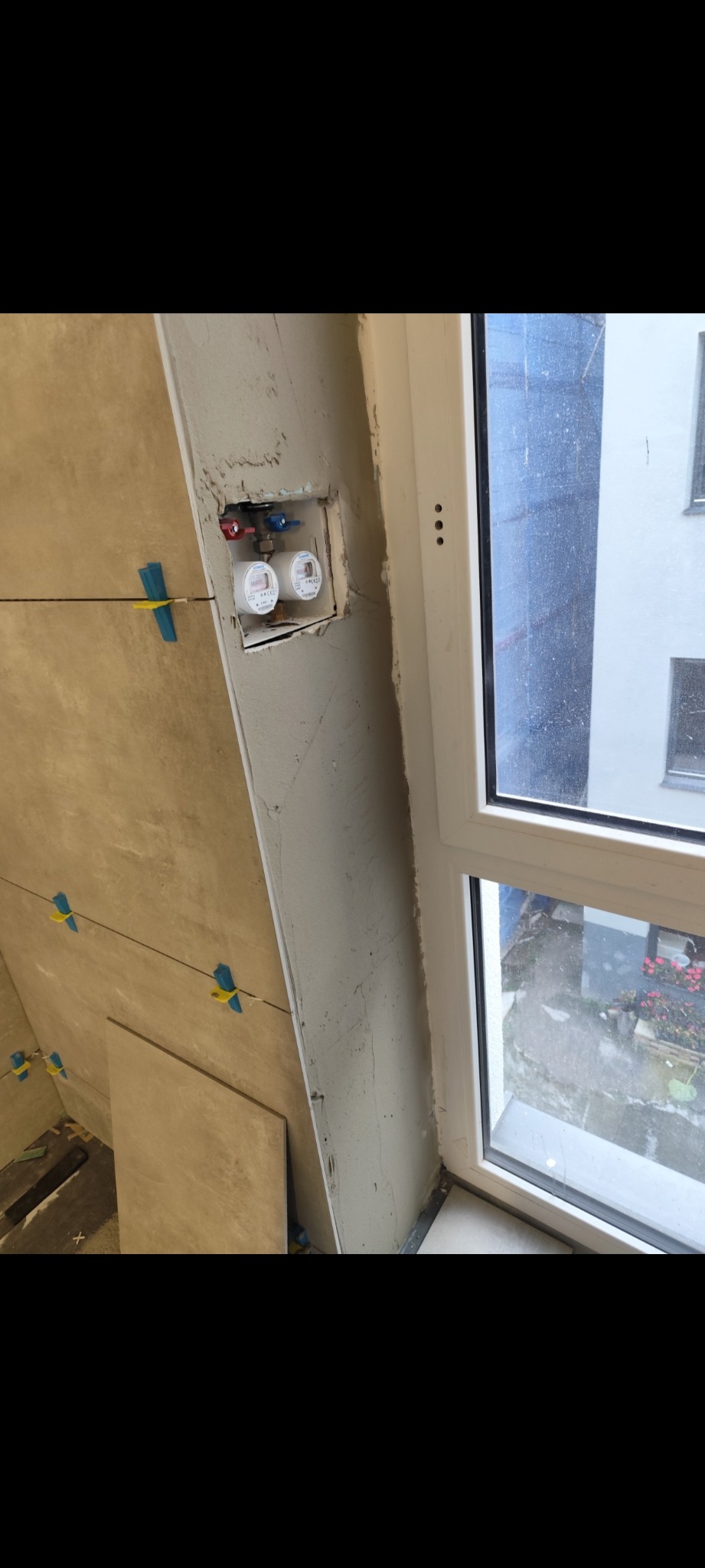 Układanie płytek przy oknie, widoczne liczniki wody i poziomowanie. Prace glazurnicze w trakcie wykańczania łazienki.