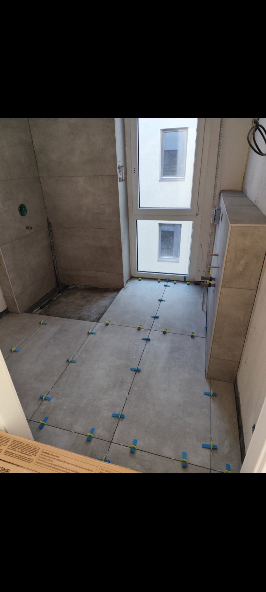 Układanie szarych płytek podłogowych w łazience, widoczne niebieskie krzyżyki dystansowe, fragment okna i karton z instrukcją montażu w rogu kadru.