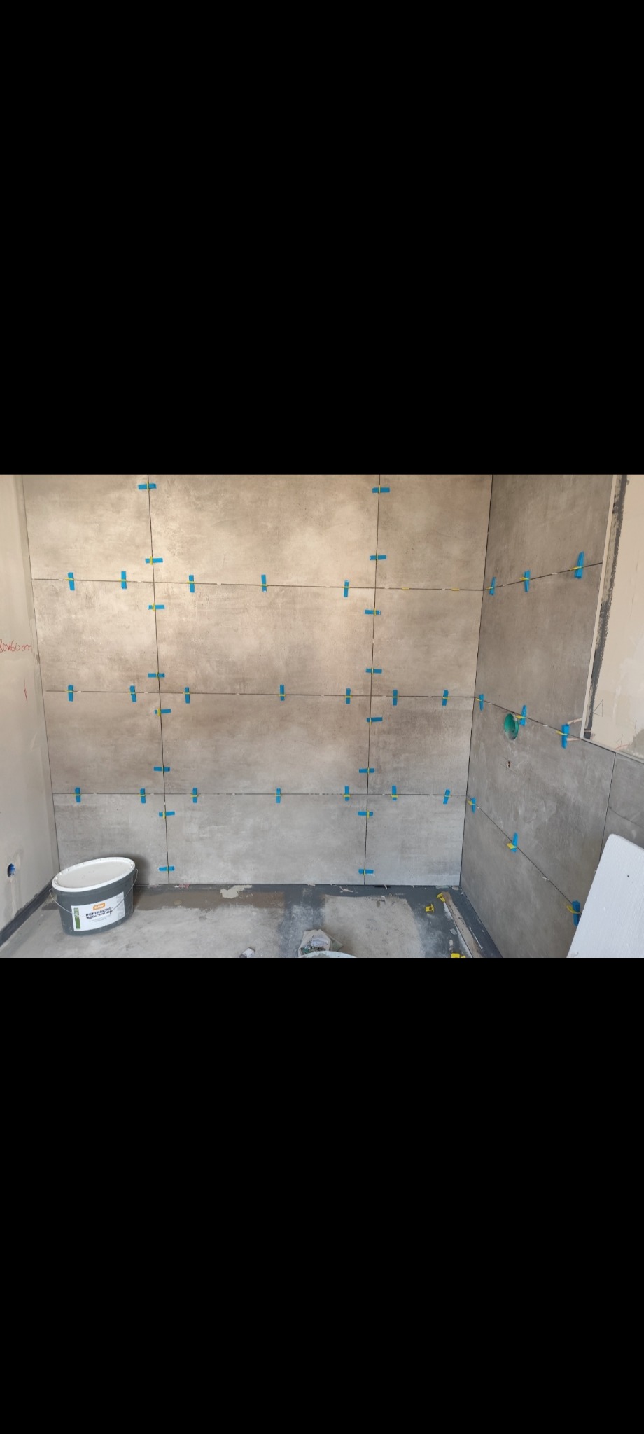 Ściana w trakcie układania płytek imitujących beton, z użyciem niebieskich krzyżyków dystansowych. Na podłodze wiadro z klejem i resztki materiałów budowlanych.