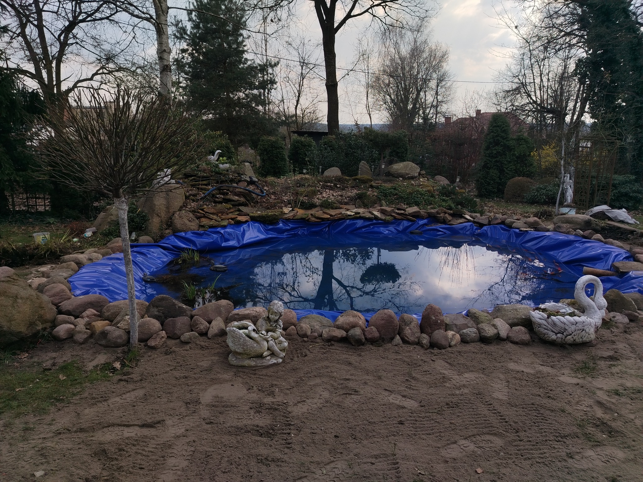 Niebieska folia w stawie ogrodowym otoczonym kamieniami i rzeźbami, odbicie drzew w wodzie. Etap budowy oczka wodnego w ogrodzie.