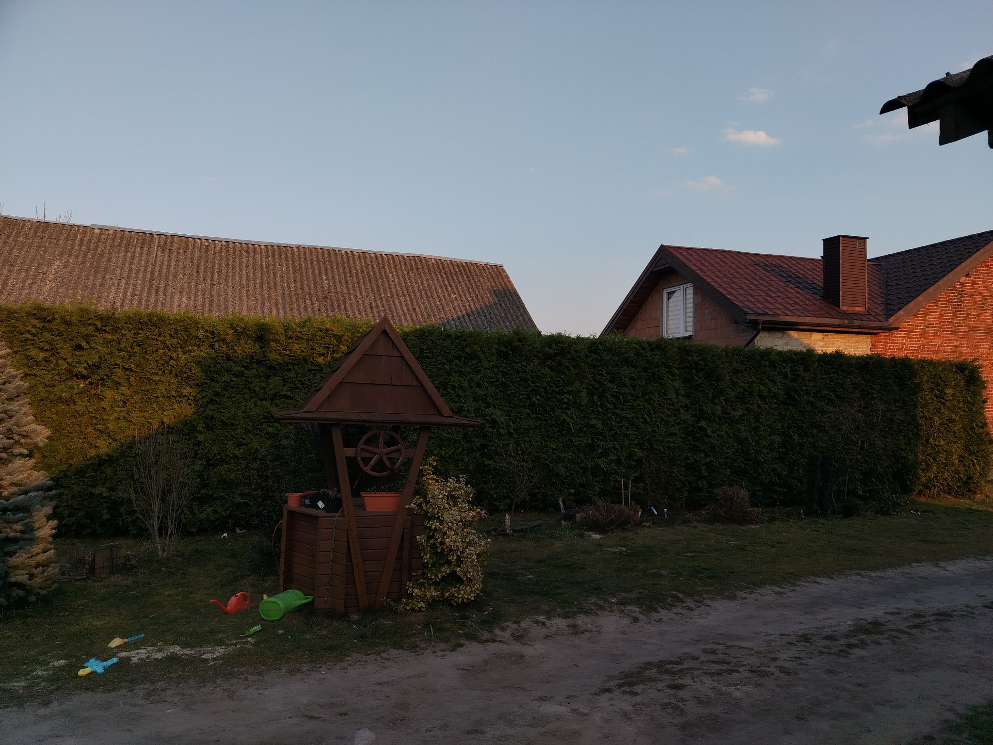 Ogród z drewnianą studnią dekoracyjną i gęstym żywopłotem w tle, wczesnym wieczorem. Dziecięce zabawki leżą na ziemi, dodając scenerii naturalnego uroku.