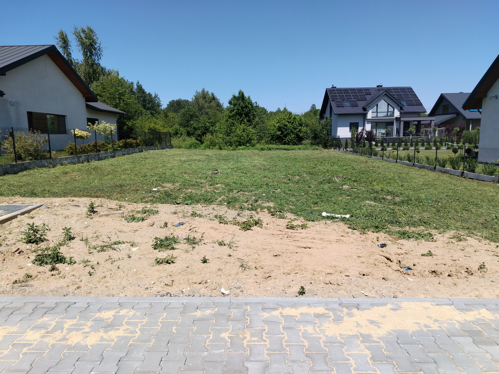 Teren przed domem z fragmentem ułożonej kostki brukowej. Widoczne trawa, piasek i nowoczesne domy w tle, słoneczny dzień.