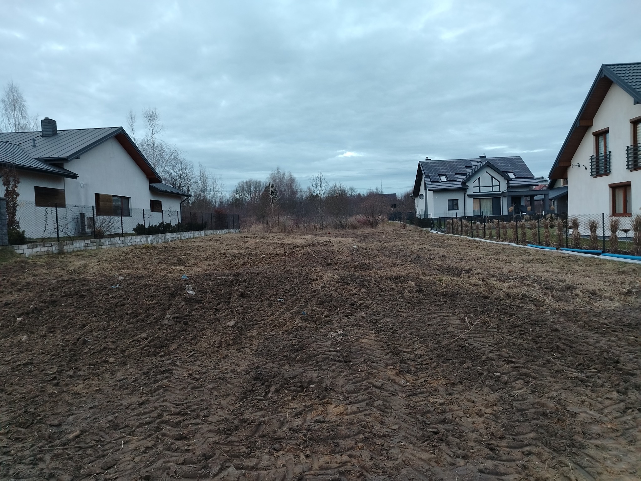 Przygotowany teren pod budowę domu z widocznymi śladami opon, w tle dwa domy jednorodzinne i pochmurne niebo. Widok na działkę budowlaną.