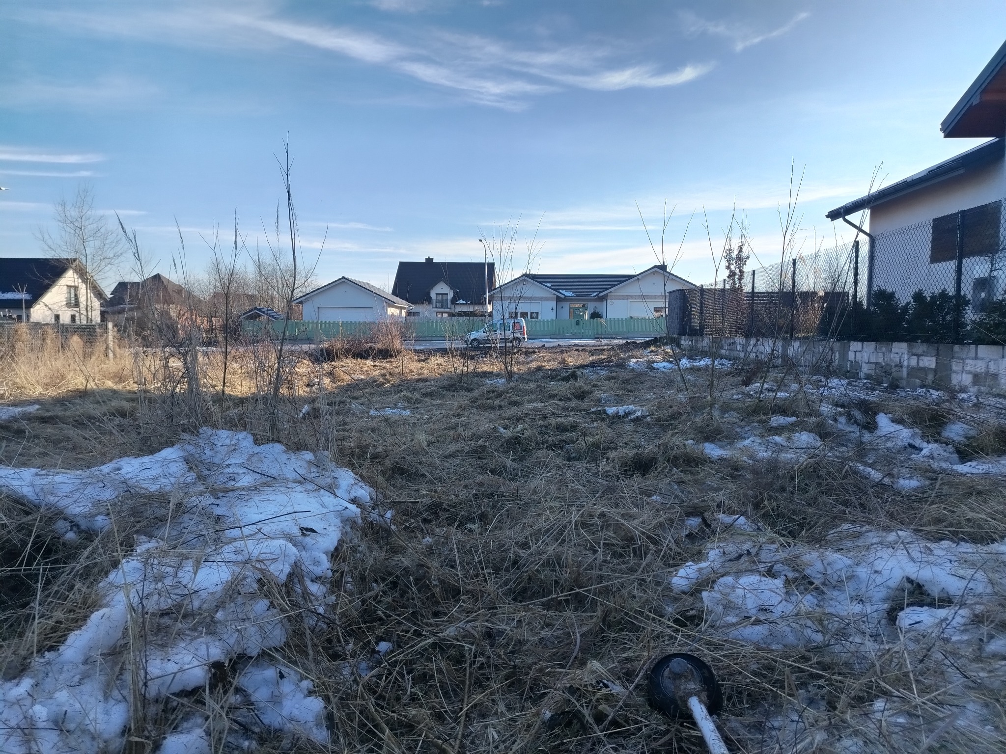 Zaniedbany teren z resztkami śniegu i suchą trawą, w tle zabudowa jednorodzinna i błękitne niebo. W dolnym rogu widoczny fragment narzędzia ogrodniczego.