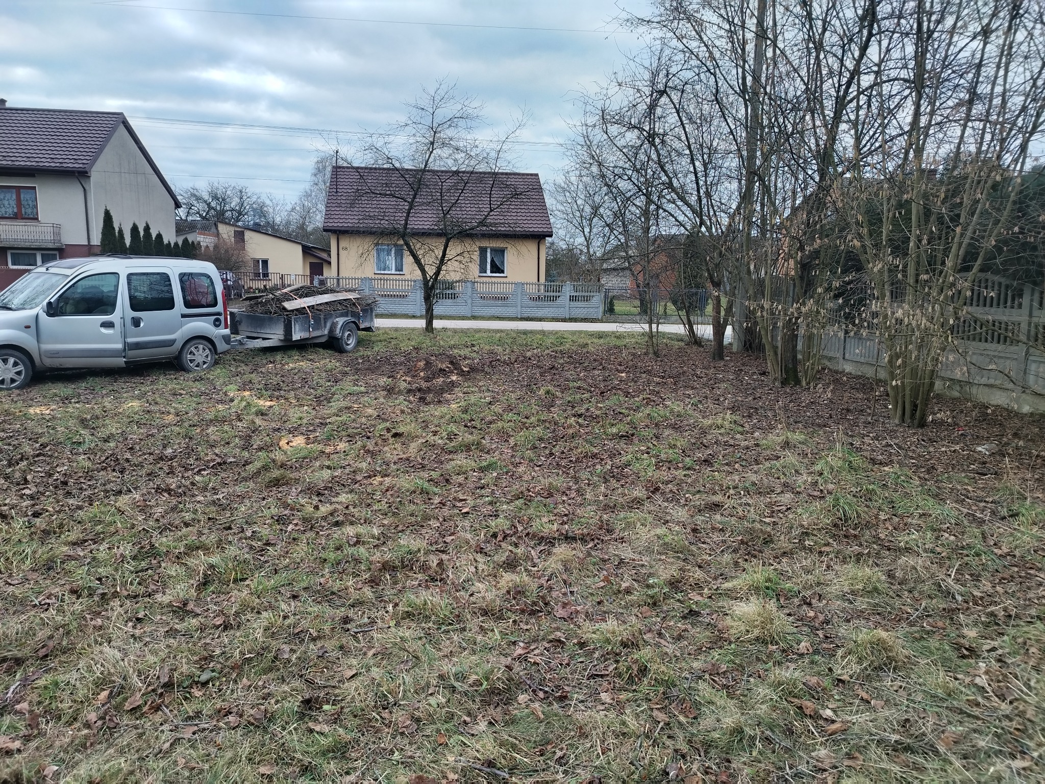 Przygotowany teren po wycince drzew, z przyczepą pełną gałęzi i pni. W tle domy jednorodzinne i pochmurne niebo.