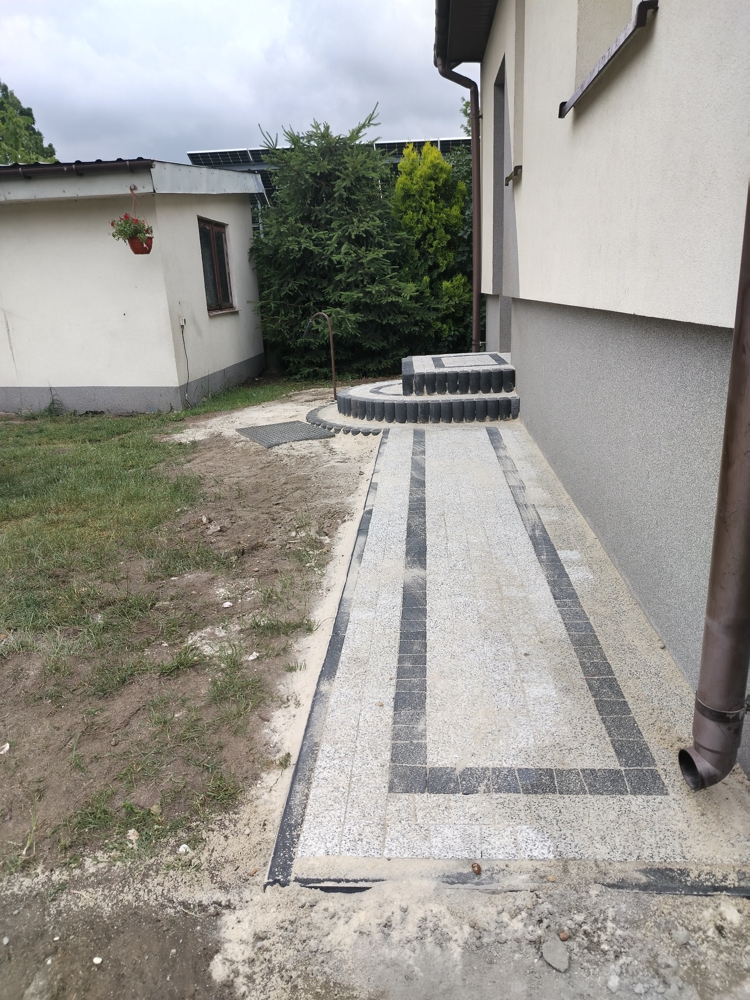 Ścieżka z kostki brukowej w odcieniach szarości prowadząca do schodów wejściowych budynku, z widocznym fragmentem elewacji i zieleni w tle.