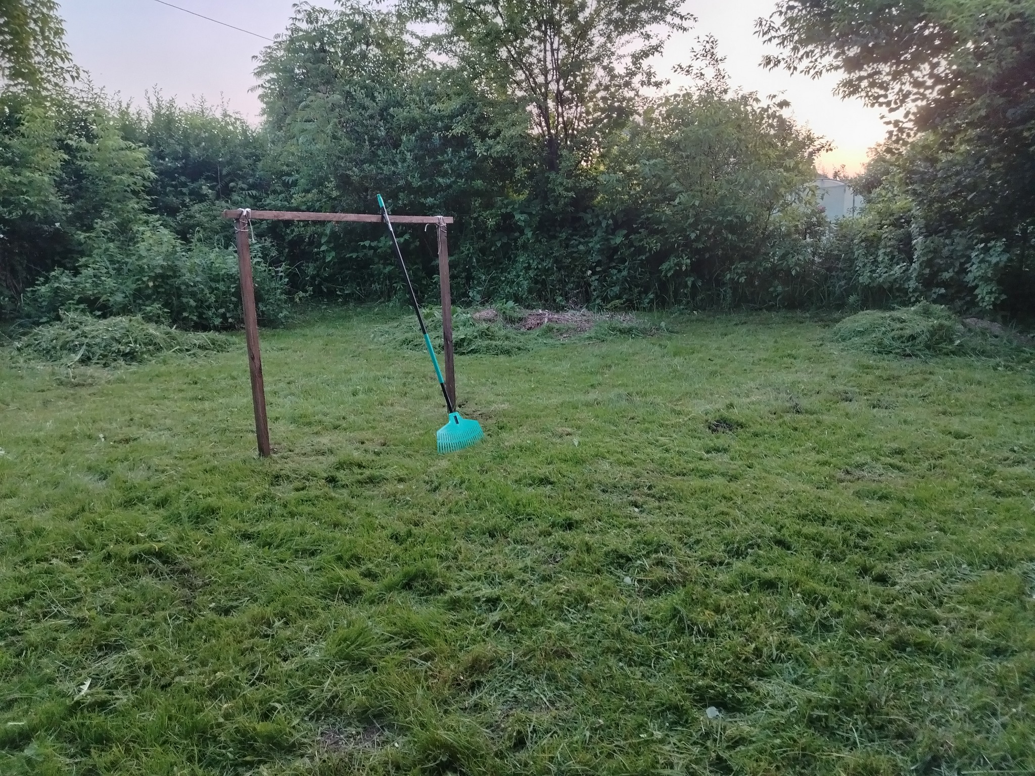 Świeżo skoszony trawnik z pozostawioną grabiami opartymi o drewnianą konstrukcję. W tle zieleń drzew i krzewów.