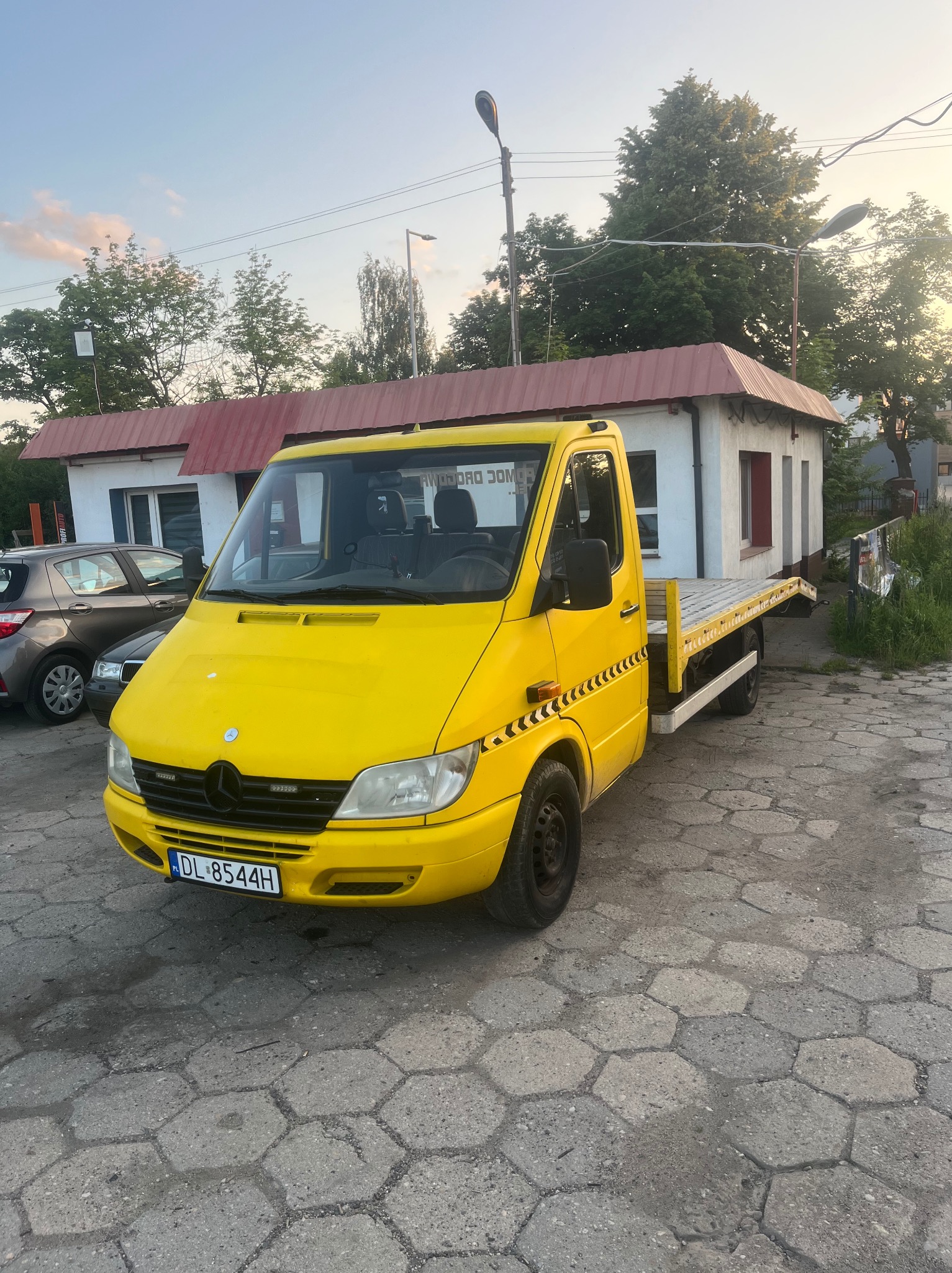 Żółta laweta Mercedes-Benz Sprinter z tablicą rejestracyjną DL 8544H, zaparkowana na brukowanym placu. W tle budynek z czerwonym dachem i drzewa.