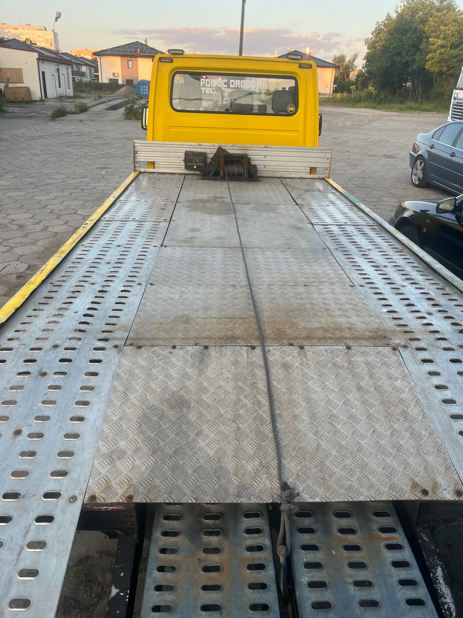 Żółta laweta z wyciągarką na platformie, widok z tyłu. Na haku lina stalowa. W tle budynki i drzewa. Platforma lawety z blachy ryflowanej, z otworami.