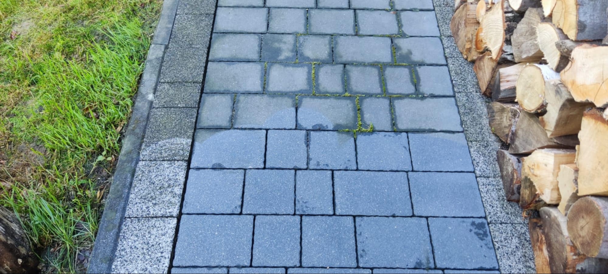 Szara kostka brukowa z widocznym mchem w spoinach, obok trawa i ułożone drewno. Kostka częściowo mokra, sugerująca niedawne mycie. Widok z góry.