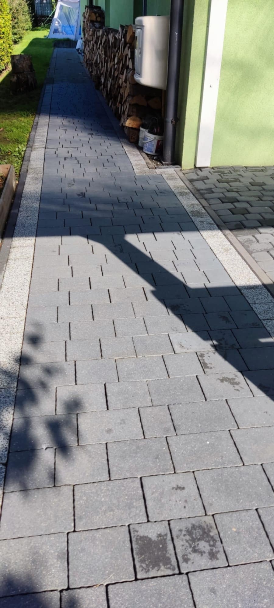 Umyta kostka brukowa na posesji. Widoczne plamy, resztki cienia drzewa. W tle zielona ściana budynku i zgromadzone drewno.