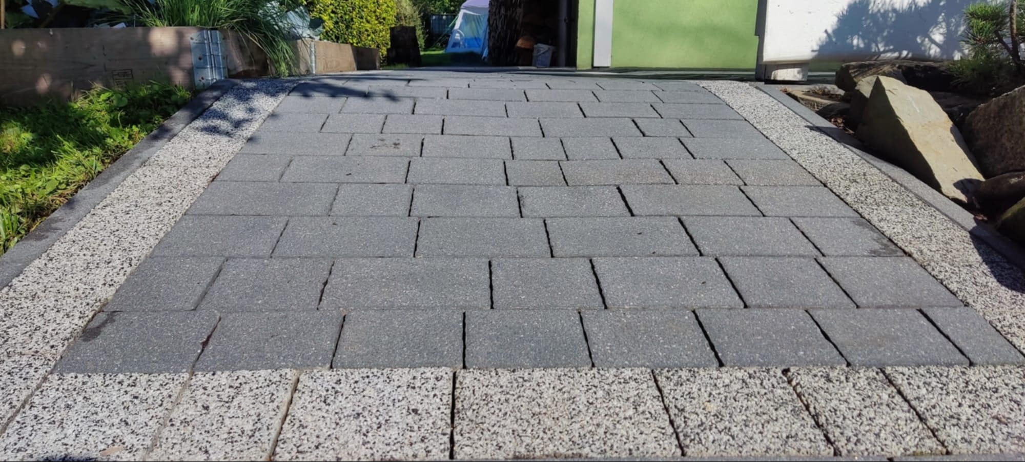 Szara kostka brukowa ułożona w regularny wzór, obramowana drobnym, jasnym żwirem. Widoczne rośliny i fragment budynku w tle. Czysta powierzchnia, symetryczny układ.