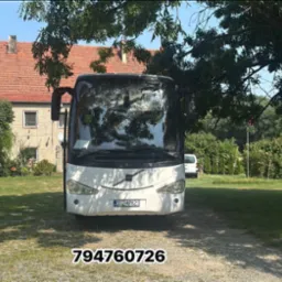 Biały autokar Volvo z numerem rejestracyjnym DDZ 47821 stoi na tle zieleni i budynku z czerwoną dachówką, częściowo zasłonięty przez gałęzie drzewa.