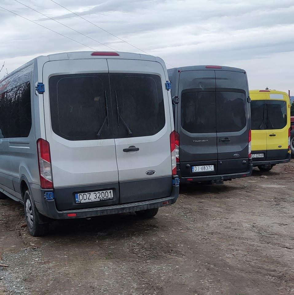 Flota trzech busów Ford Transit (srebrny, szary, żółty) zaparkowana na ziemistym placu, widok z tyłu, tablice rejestracyjne widoczne. Pochmurne niebo w tle.