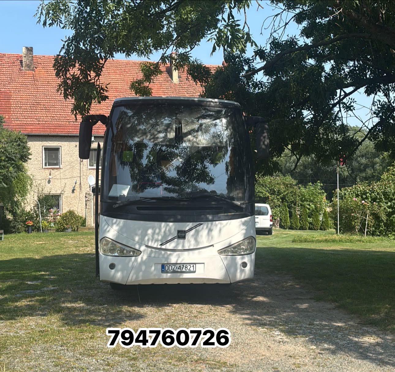 Biały autokar Volvo z numerem rejestracyjnym DDZ 47821 stoi na tle zieleni i budynku z czerwoną dachówką, częściowo zasłonięty przez gałęzie drzewa.